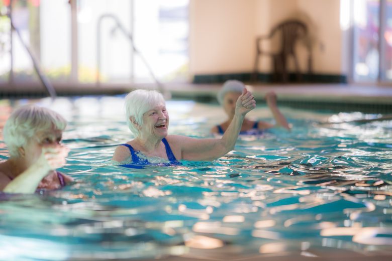 Three older women enjoy water aerobics in an indoor pool, smiling and raising their arms as sunlight streams through large windows in the background.