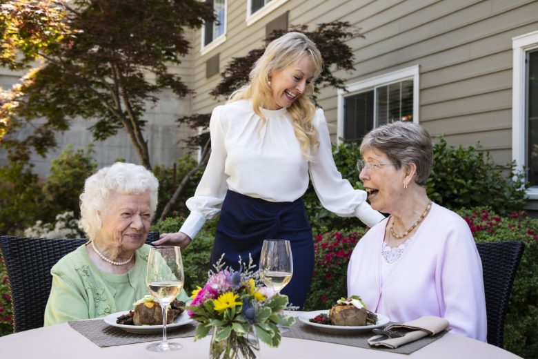 Two older women sit at an outdoor table with meals and wine, smiling and talking. A younger woman stands between them, also smiling. There are flowers on the table and a garden with greenery in the background.