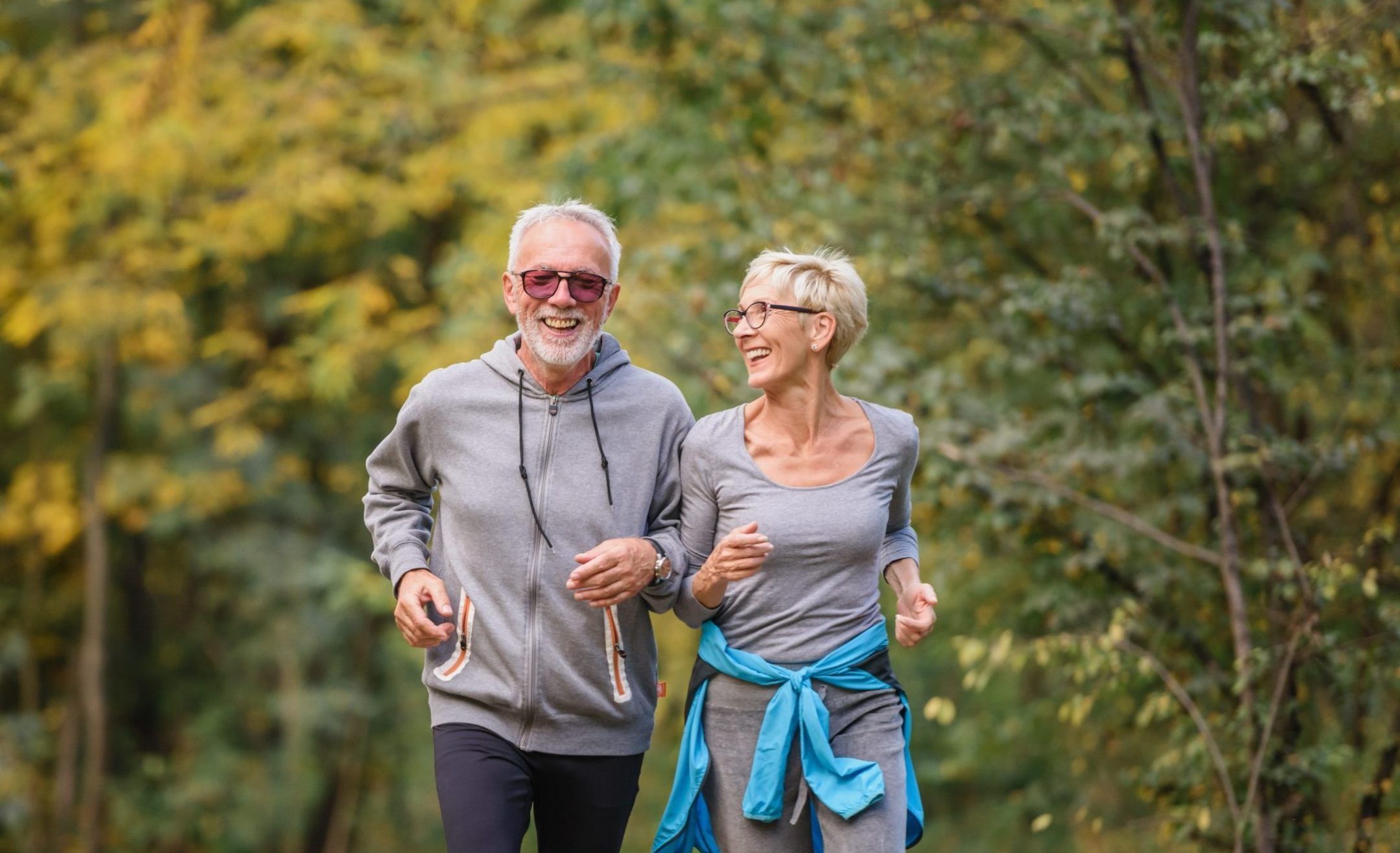 An older man and woman jog outdoors on a wooded trail, smiling in casual athletic clothes. Early autumn trees form a green and yellow backdrop, reflecting the active and vibrant Eskaton Village Placerville lifestyle.