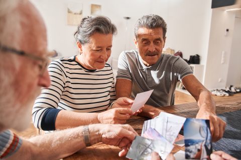 Three older adults sit at a table indoors at Eskaton Village Placerville memory care, warmly sharing printed photographs and reminiscing together in a nostalgic atmosphere.