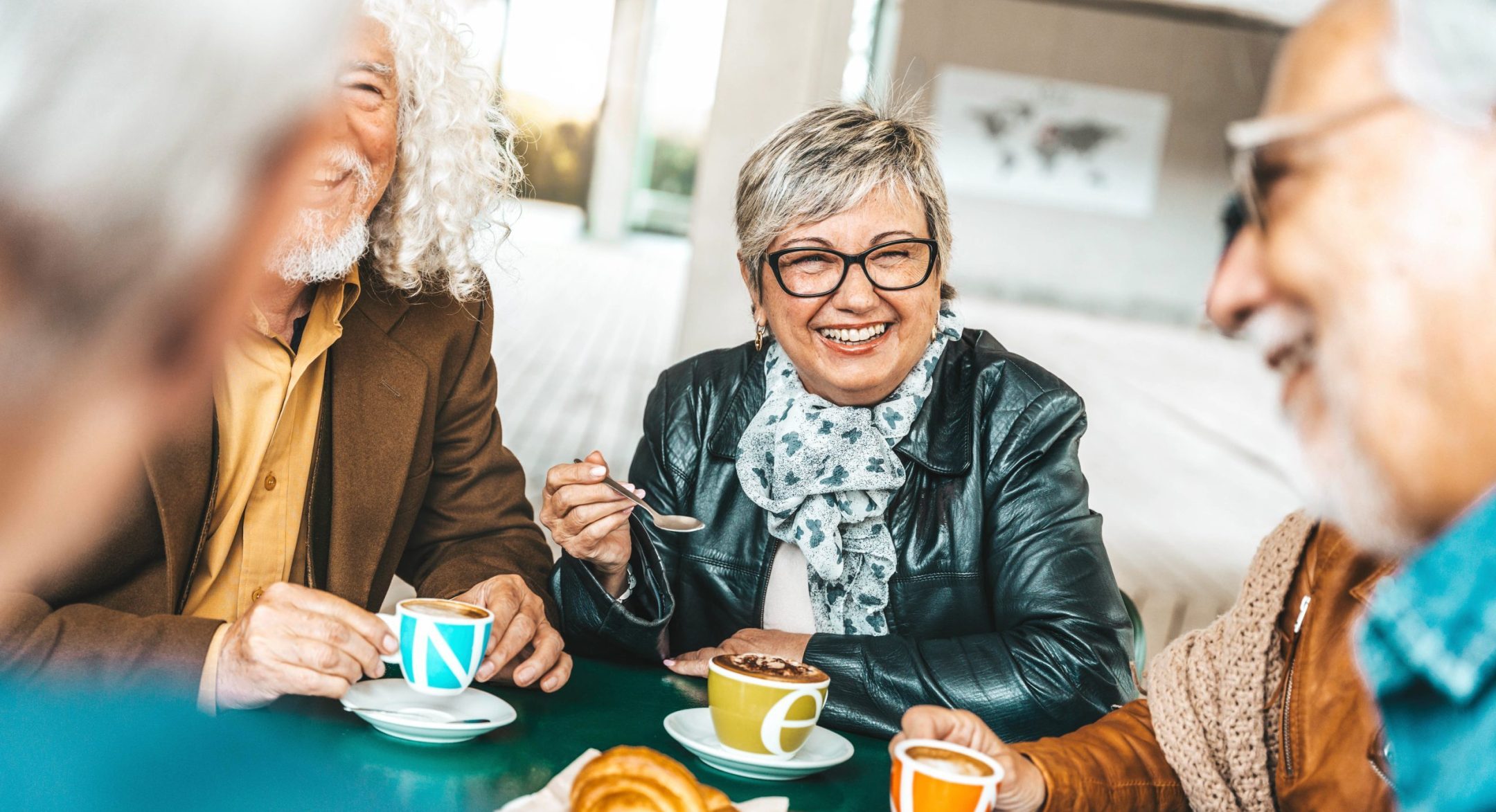 A group of older adults sit together at a table, smiling and enjoying coffee, hot chocolate, and pastries. One woman in glasses and a scarf holds a spoon, looking cheerful as she talks with friends.