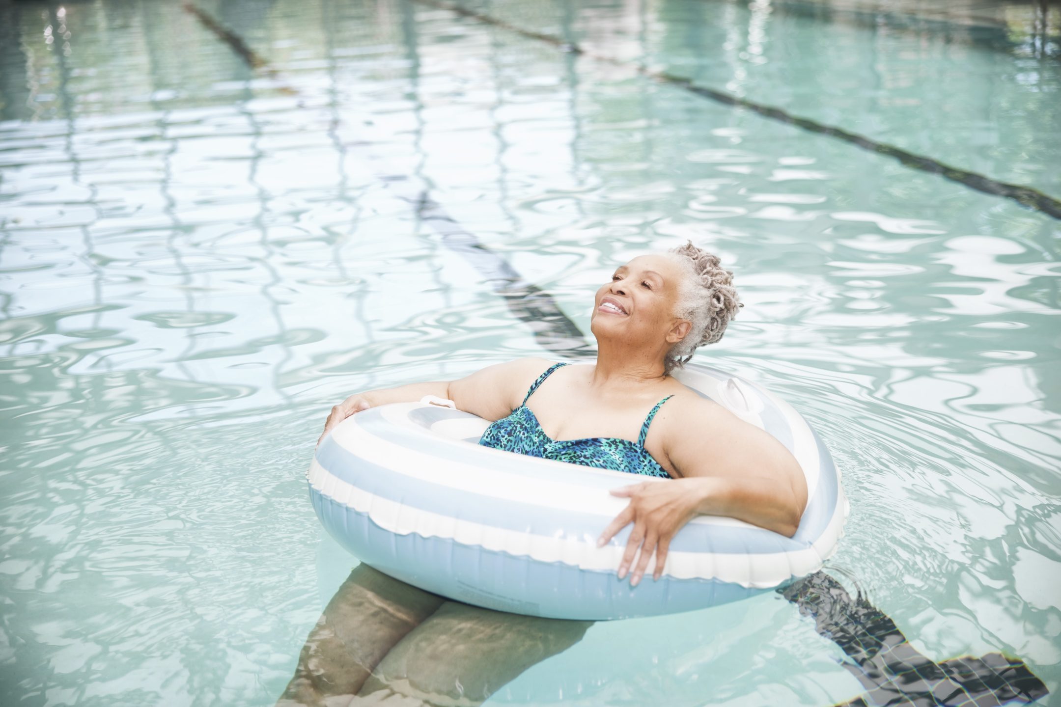 An Eskaton Village Placerville resident enjoys a relaxing experience, smiling as she floats in a clear swimming pool with a white and blue inflatable ring, surrounded by inviting swimming lanes.