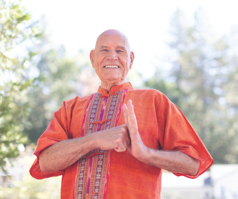 An older man in an orange traditional shirt stands outdoors at Eskaton Village Placerville, smiling warmly with his hands pressed together in greeting. Trees and soft sunlight highlight the welcoming lifestyle in the background.
