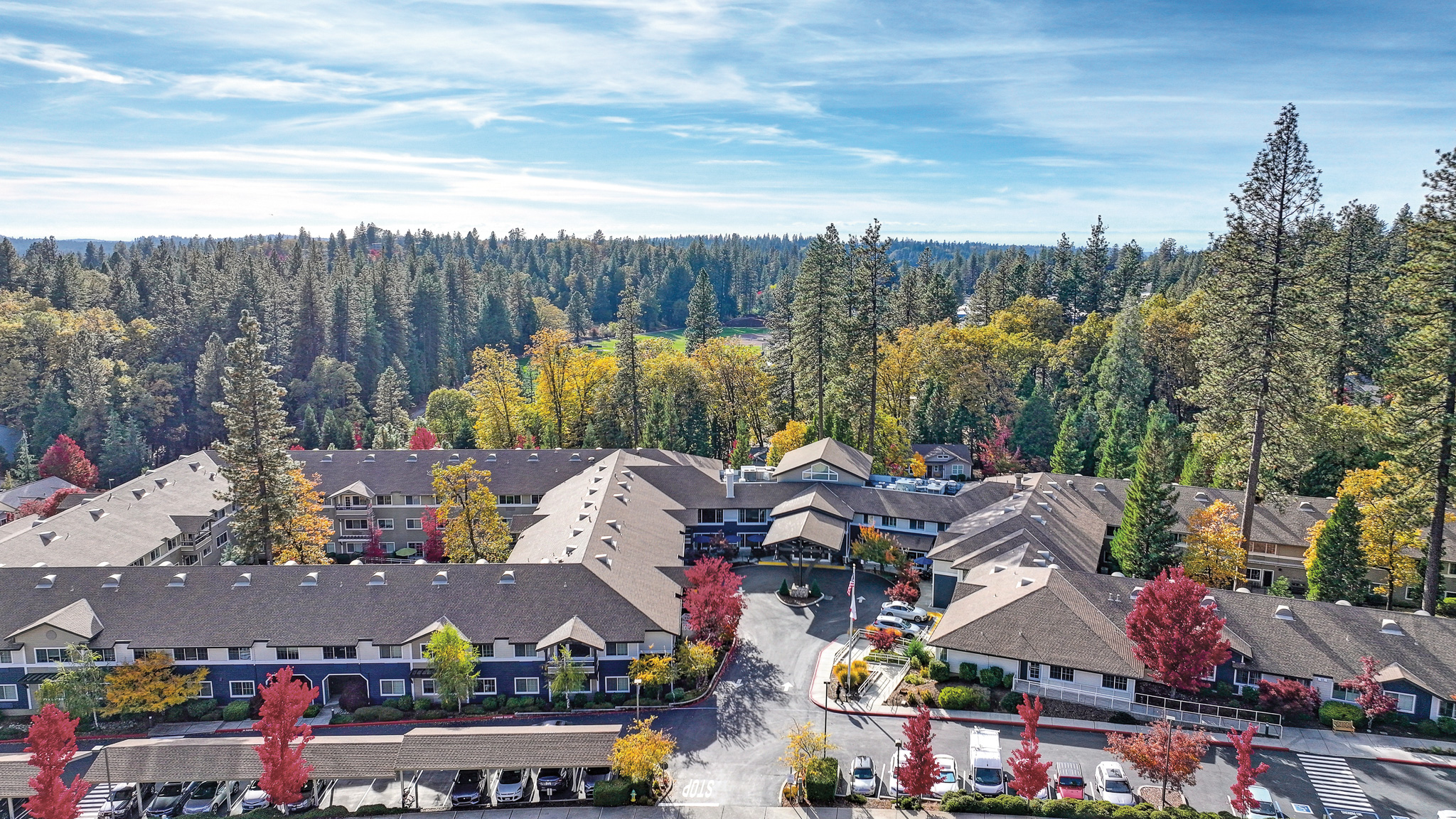 Aerial view of Eskaton Village Grass Valley, a large multi-wing building surrounded by autumn trees, parked cars, and a clear sky above the forested landscape.
