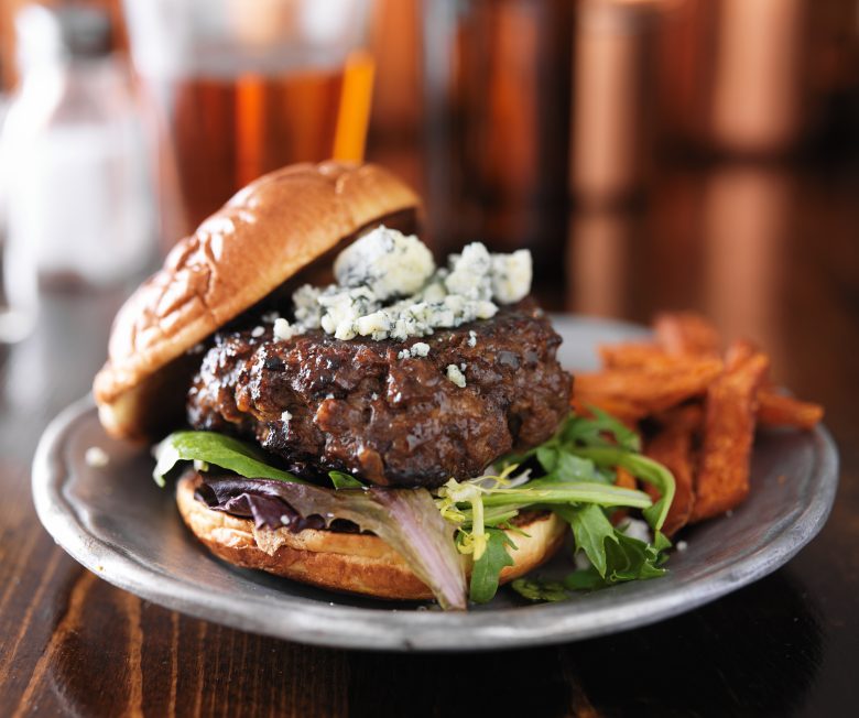 A close-up of a gourmet burger with blue cheese crumbles, leafy greens, and a toasted bun, served on a metal plate with sweet potato fries in the background, highlights the delicious offerings at Eskaton Grass Valley dining.