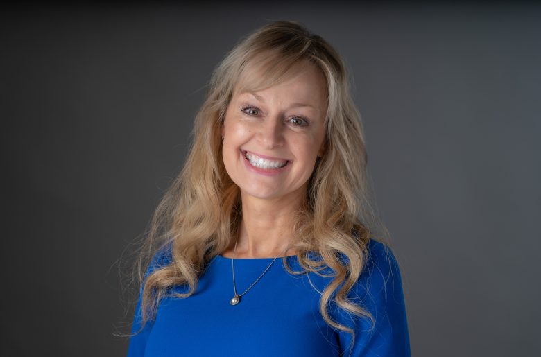 A smiling woman with long, wavy blonde hair, wearing a blue top and delicate pendant necklace, poses in front of a plain gray background at Eskaton Village Grass Valley senior living.