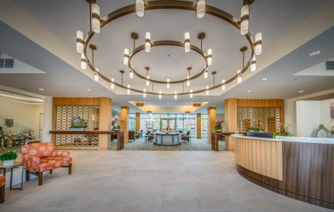 Spacious, modern hotel lobby with a large circular chandelier, contemporary seating area, reception desk, decorative plants, and large windows letting in natural light.