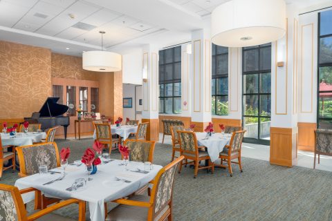 Elegant dining room with round tables set with white tablecloths, folded red napkins, and wooden chairs. Large windows let in natural light, and a grand piano sits in the corner near patterned wallpaper.