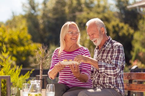 An elderly couple sits on a bench outdoors, smiling and sharing food from a plate. The woman wears a striped shirt and glasses, while the man wears a plaid shirt. Sunlight and greenery surround them, creating a cheerful atmosphere.