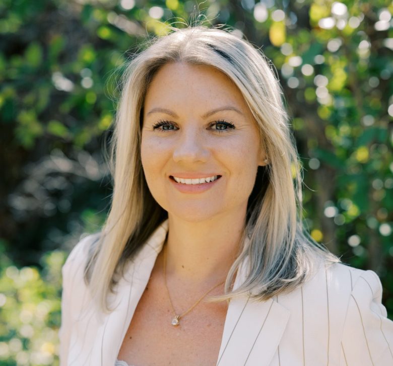 A woman with straight, shoulder-length blonde hair and a white blazer smiles outdoors. The background is filled with green foliage and dappled sunlight.