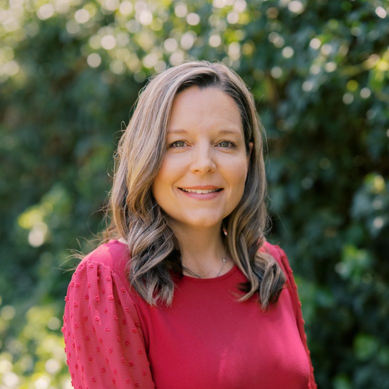 A woman with wavy, light brown hair wearing a red textured long-sleeve top smiles outdoors, with green foliage softly blurred in the background.
