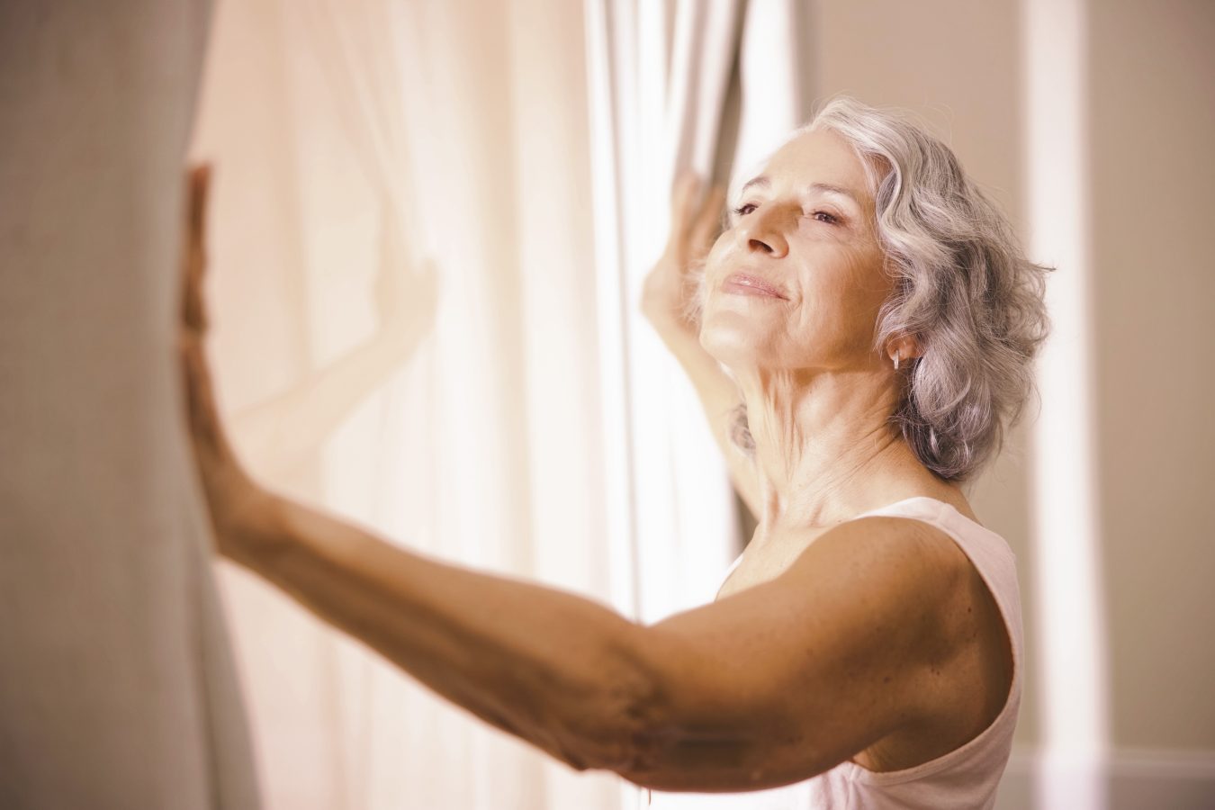 An older woman with gray hair smiles softly as she opens light-colored curtains, letting in sunlight. She appears peaceful and content, standing in a sunlit room.