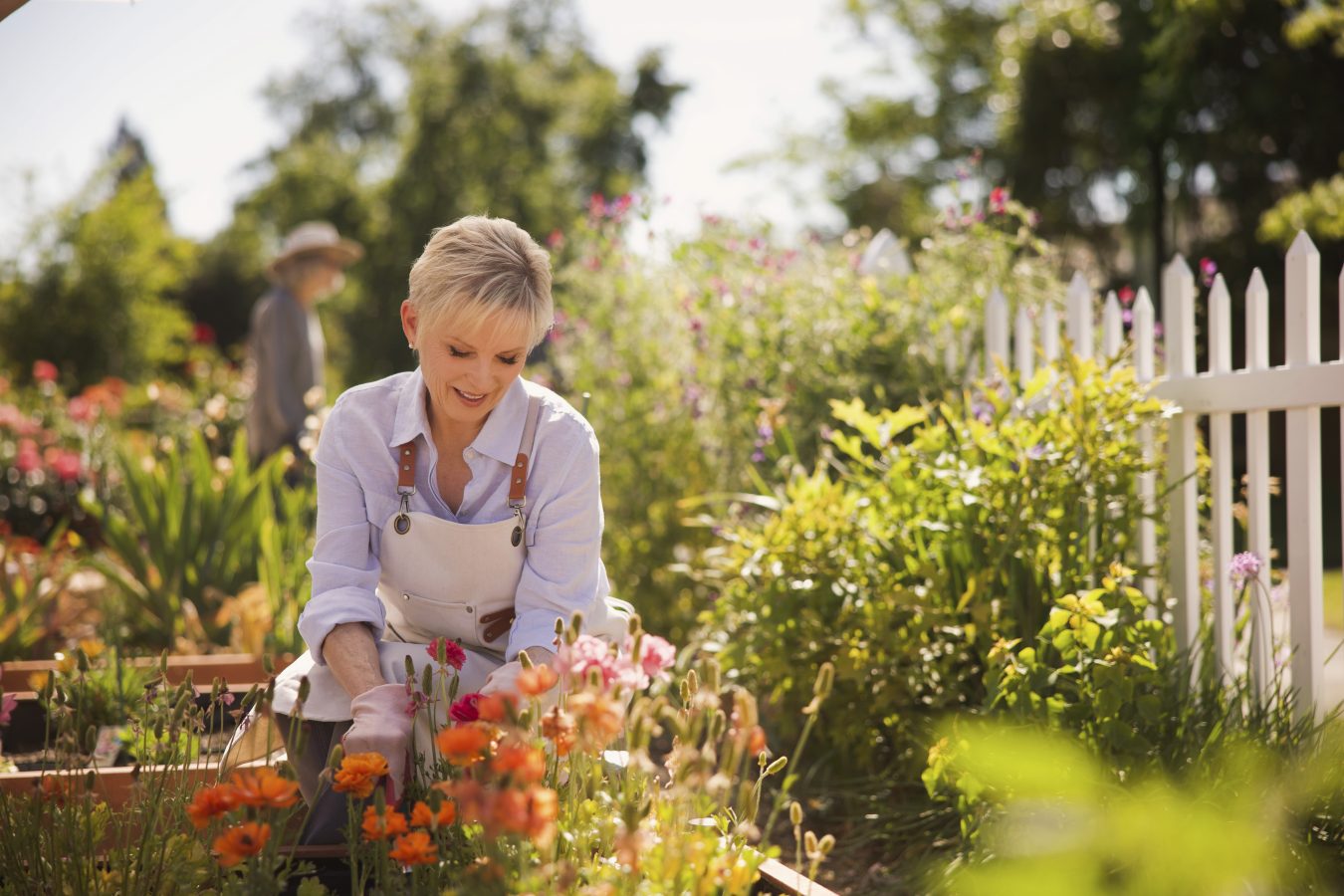 A smiling woman wearing a white shirt and apron tends to colorful flowers in a bright garden, with green plants and a white picket fence in the background. Another person is slightly blurred in the distance.