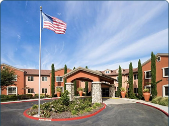 A large building with red and beige walls and tall windows, featuring a covered entrance supported by stone columns. An American flag on a tall pole waves in front, with landscaping and a circular driveway.