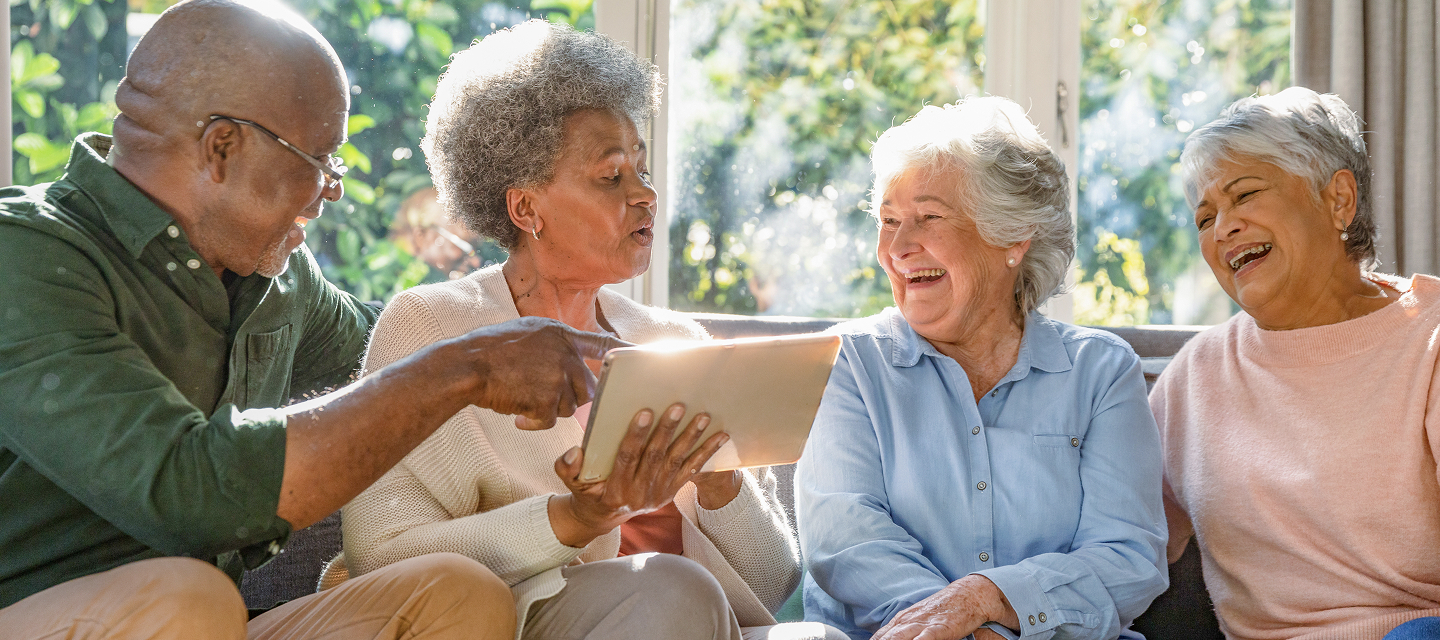 Four older adults sit together on a couch, smiling and laughing while looking at a tablet. Sunlight streams through a window behind them, creating a warm and cheerful atmosphere.