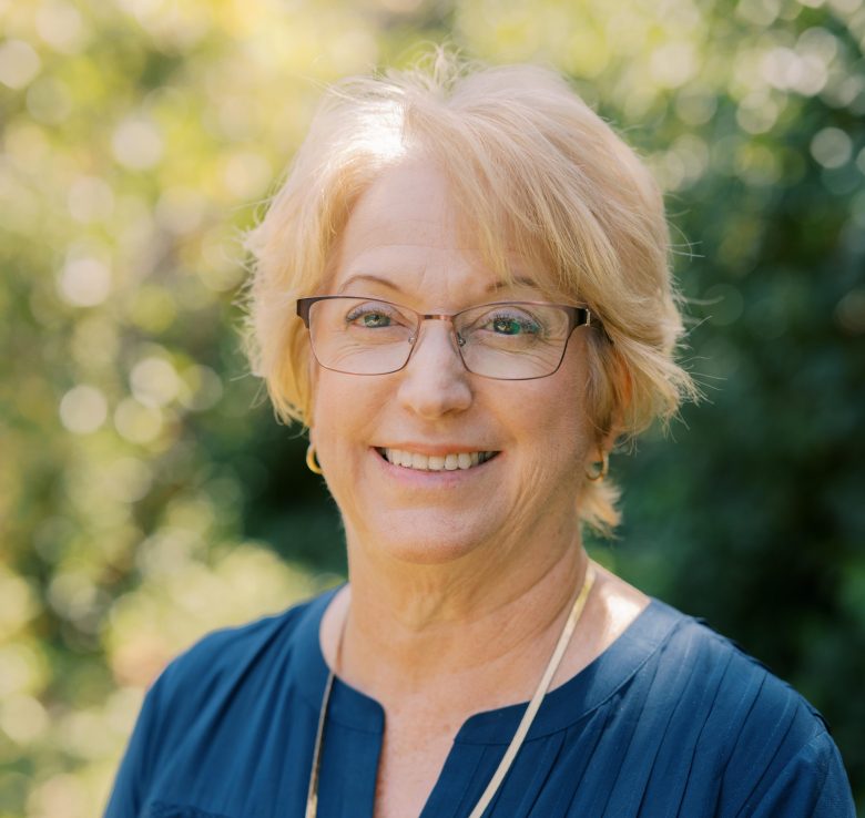 A middle-aged woman with short blonde hair, glasses, and a blue blouse smiles outdoors with greenery and sunlight blurred in the background.