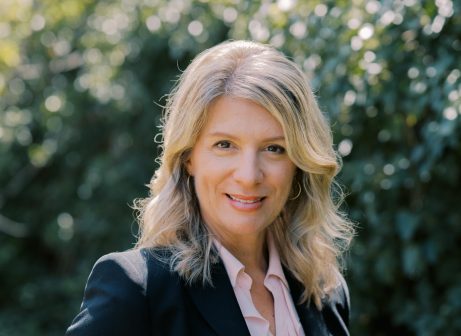 A woman with blonde hair wearing a black blazer and light pink blouse stands outdoors, smiling, with greenery and sunlight blurred in the background.