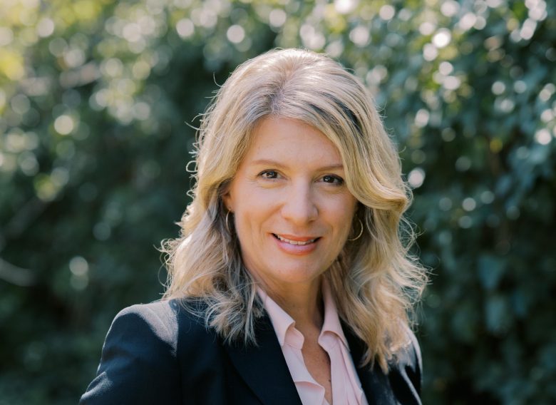A woman with blonde hair wearing a black blazer and light pink blouse stands outdoors, smiling, with greenery and sunlight blurred in the background.