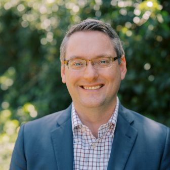 A man wearing glasses, a blue blazer, and a checkered shirt smiles outdoors with greenery and sunlight in the blurred background.