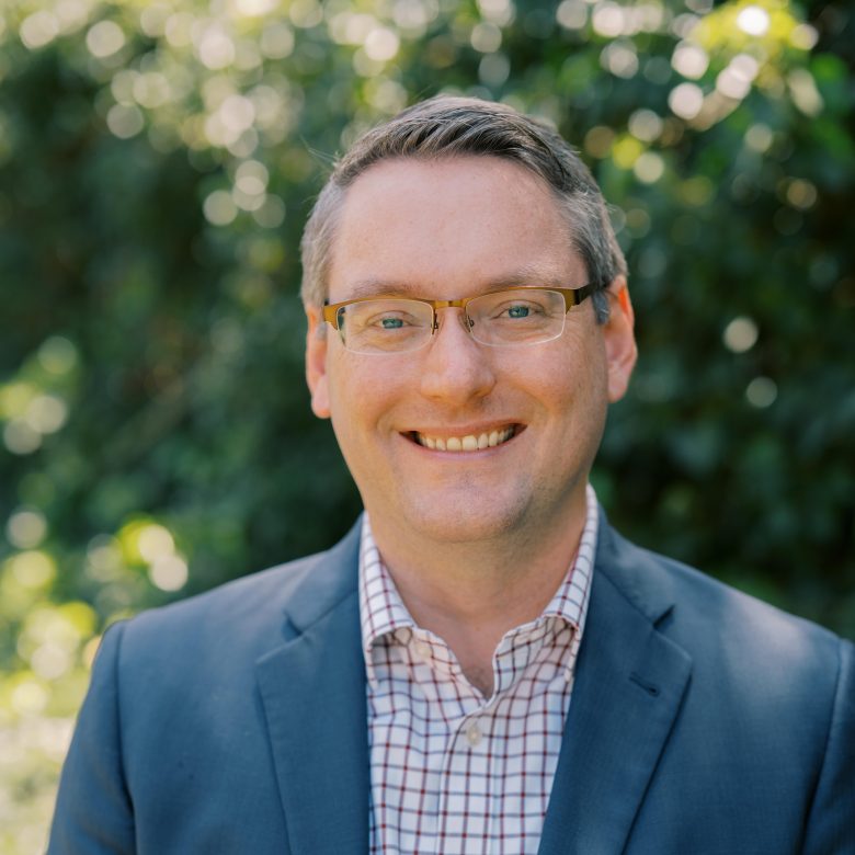 A man wearing glasses, a blue blazer, and a checkered shirt smiles outdoors with greenery and sunlight in the blurred background.