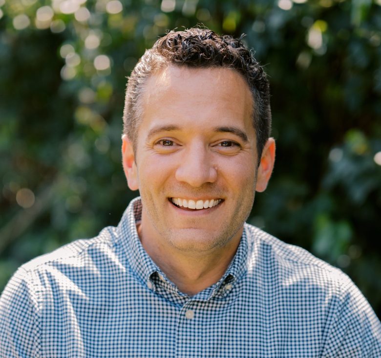 A man with short curly hair smiles outdoors, wearing a blue checkered button-up shirt. Green foliage and sunlight create a blurred natural background.