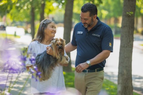 An older woman holding a small dog smiles at a man in a navy polo shirt with a name tag as they walk together on a tree-lined path. The scene is sunny and peaceful.