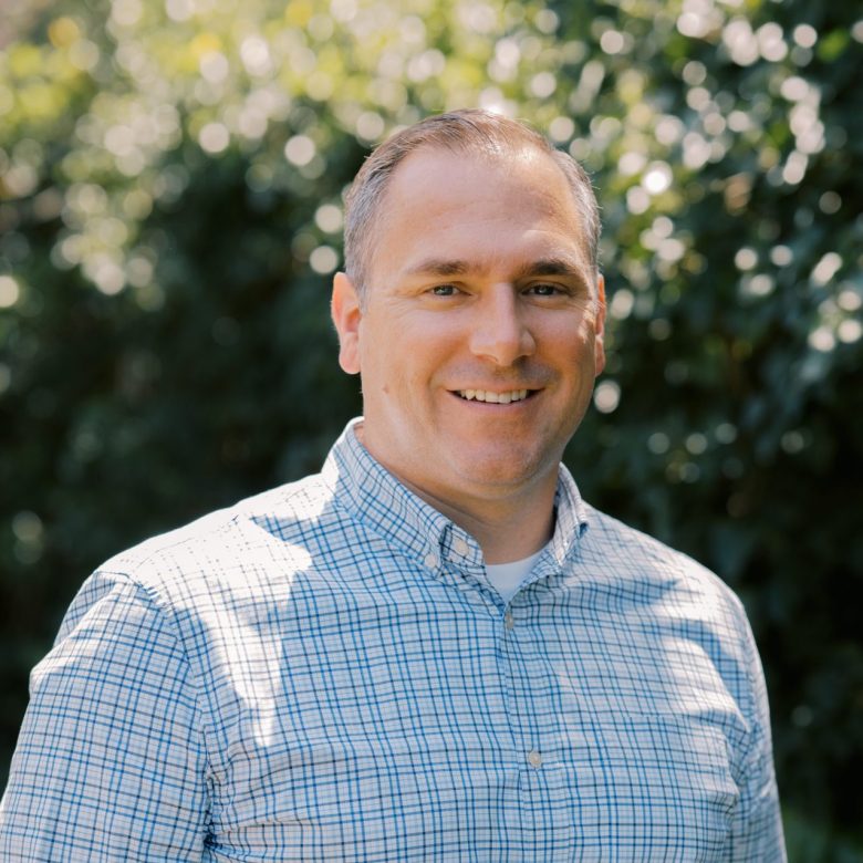 A man wearing a blue and white plaid shirt smiles while standing outdoors, with sunlight and green leafy trees blurred in the background.