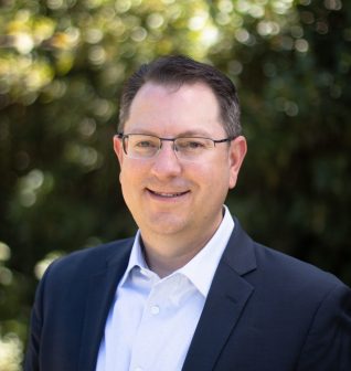 A man with short brown hair, glasses, and a navy blazer over a white shirt smiles outdoors, with greenery and sunlight softly blurred in the background.