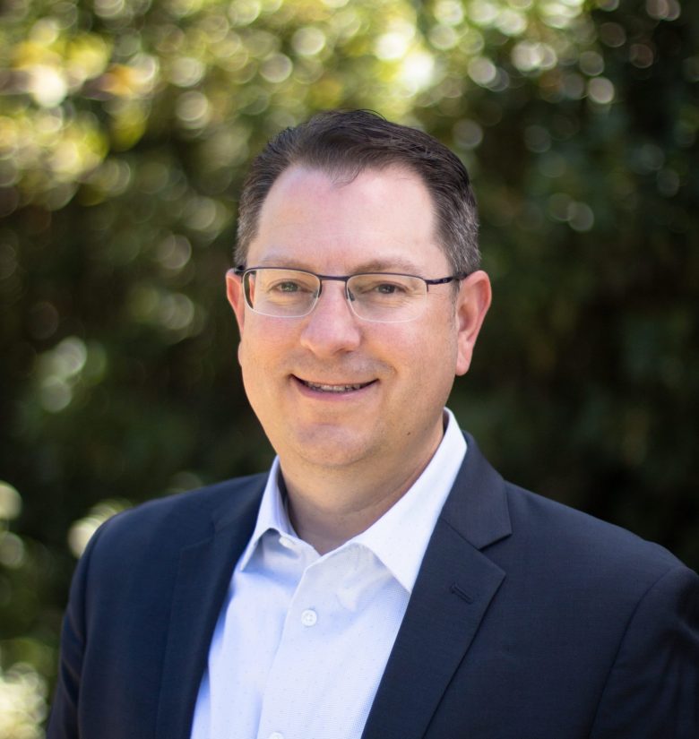 A man with short brown hair, glasses, and a navy blazer over a white shirt smiles outdoors, with greenery and sunlight softly blurred in the background.