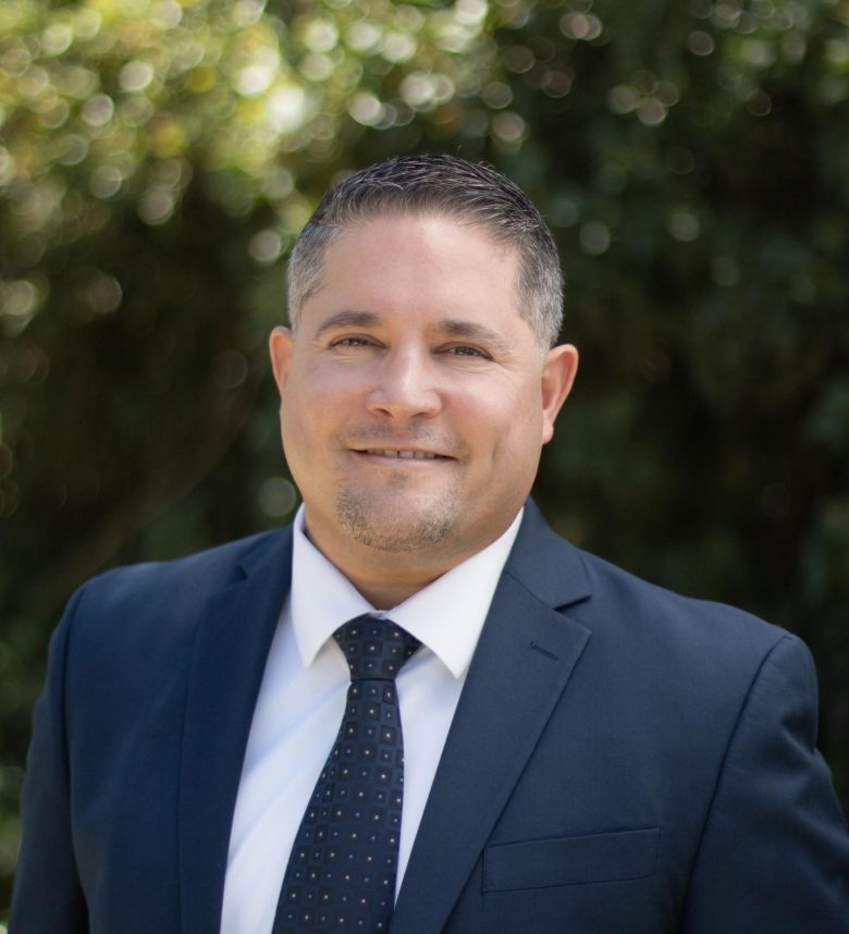 A man in a dark suit and white shirt with a patterned tie smiles at the camera, exuding leadership as he stands outdoors with blurred green foliage in the background.