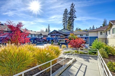 A sunny day at the modern Eskaton residential complex with blue buildings, colorful trees, bright landscaping, and a ramp leading to the main entrance under a wooden canopy. Tall pine trees frame the clear blue sky in the background.